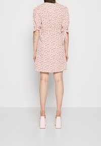 Kvinna klädd i en vit kort blommig klänning med puffärmar knutna vid armbågarna, tillsammans med ljusrosa klackskor med blockklack, stående med ryggen mot kameran.