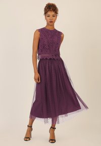 Abito di pizzo viola con design senza maniche, dettagli intricati e gonna in tulle a strati con pois, abbinato a sandali neri con cinturini.