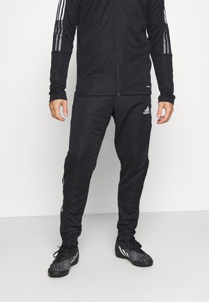 Mężczyzna w czarnych spodniach dresowych Adidas, czarnej kurtce na zamek z paskami na rękawach oraz czarnych butach sportowych z białymi akcentami.