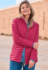 Femme souriante portant une veste matelassée rouge sur une chemise en denim bleu et un jean, se tenant à l'extérieur contre un mur en pierre.