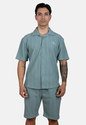Hombre con cabello corto y oscuro y tatuajes lleva una camisa de manga corta azul verdoso con botones y pantalones cortos a juego sobre un fondo claro y liso.