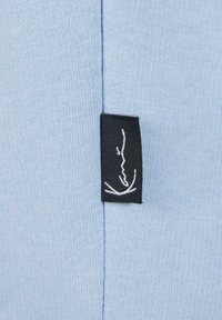 Gaiši zils audums ar gludu tekstūru un šuvēm, ar mazu melnu etiķeti ar baltām šuvju detaļām.