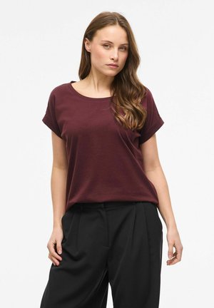 Kvinde med langt brunt hår har en løs bordeaux t-shirt og sorte bukser på, stående mod en ensfarvet hvid baggrund.