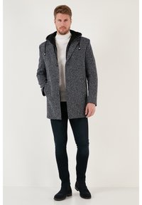 Abrigo de hombre en gris y negro con estampado, capucha, cierre de botones, dos bolsillos y un diseño texturizado tejido. Llevado sobre un cuello de tortuga.