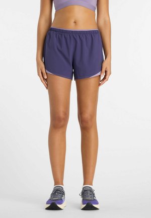 Femme portant un short de course violet, un soutien-gorge de sport assorti et des chaussures de course violettes et grises, debout sur un fond blanc.