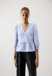 Blå og hvit stripete bluse med V-hals, puffede ermer og peplumliv, kombinert med svarte vide bukser.