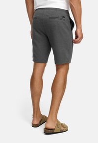 Hombre vestido con shorts gris oscuro y sandalias beige, de pie con la espalda hacia el espectador frente a un fondo blanco liso.