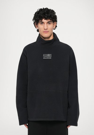 Pull noir en polaire avec col montant et logo graphique. Il présente une coupe décontractée, des manches longues et une poche avant pour plus de praticité.