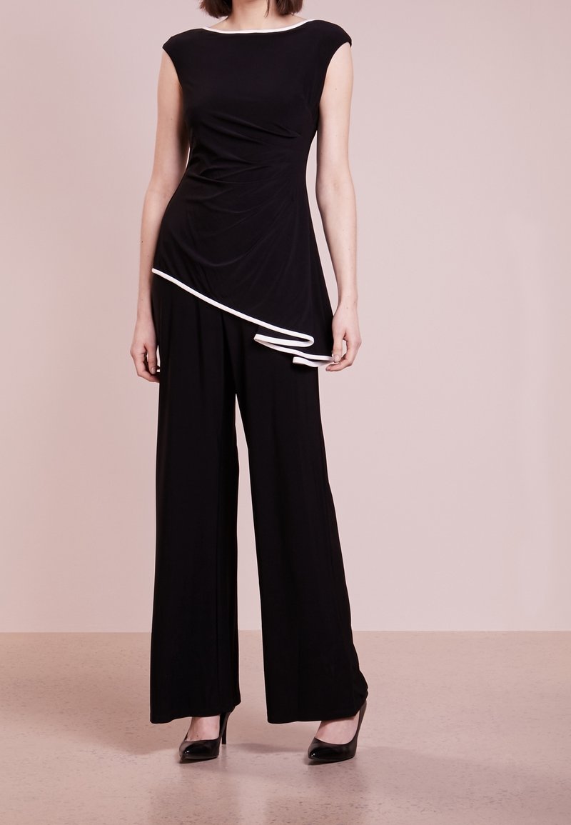 Femme portant un haut noir sans manches avec un bord blanc et un pantalon noir à jambes larges, debout sur un sol beige contre un mur rose clair.