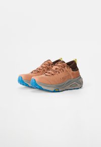 Chaussures de trail HOKA avec une tige marron et beige, présentant une semelle intermédiaire grise et une semelle extérieure Vibram bleue. Design à lacets avec une languette à l'arrière.