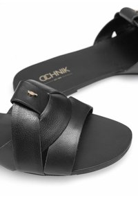 Czarne skórzane sandały z designem na krzyż, teksturowanym wykończeniem i małym złotym logo po boku; płaska podeszwa i minimalistyczny styl.