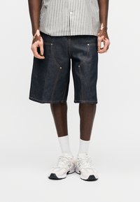 Άτομο που φοράει σκούρο τζιν σορτς μέχρι το γόνατο, λευκά αθλητικά παπούτσια, λευκές κάλτσες και ριγέ πουκάμισο, στέκεται μπροστά σε απλό φόντο.