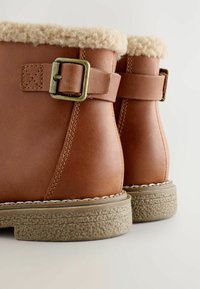 Braune Leder-Stiefel bis ankle mit cremefarbener Fleece-Fütterung, Messing-Schnallenriemen und strukturierten Gummisohlen auf einem weißen Hintergrund.