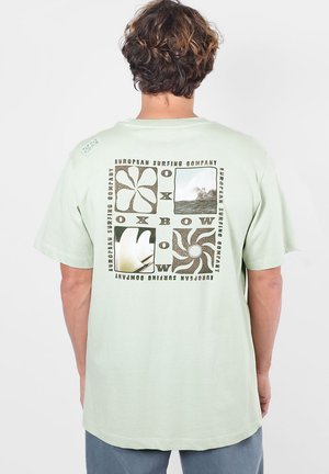 Homme portant un t-shirt vert clair avec des graphiques noirs sur le thème du surf et le texte ""European Surfing Company"" dans le dos.
