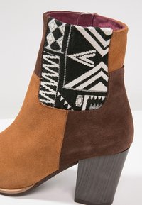 Bottine à talon carré en daim marron et beige, avec un panneau latéral en tissu à motif géométrique noir et blanc.