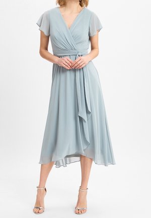 Kobieta ubrana w jasnoniebieską sukienkę midi z kopertowym dekoltem i falbanowymi rękawami oraz srebrne sandały na kostkę, stojąca na białym tle.