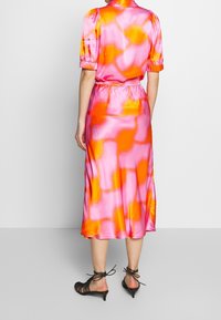 Robe midi en satin rose avec des motifs abstraits orange et jaune ; manches courtes bouffantes ; taille élastique ; fente sur le côté ; associée à des talons noirs à lanières.
