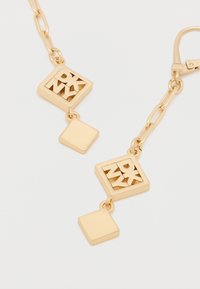 Kuldse tooniga ripptikandid, mis sisaldavad kahte geomeetrilist kujundit: ruut lõigatud tähtedega ja täisruut. Valmistatud metallist, millel on läikiv viimistlus.