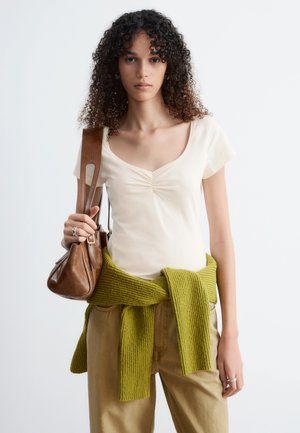 Jonge vrouw met krullend haar, gekleed in een crème kleurige top met korte mouwen, kaki broek, groene trui om de taille gestrikt en een bruine leren schoudertas.