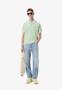Grön och vit randig polo skjorta med krage, i kombination med ljusblå raka jeans och gula sneakers, hållande en beige stickad tröja.