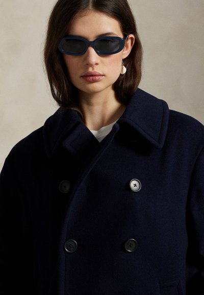 Naine, kes kannab tumedaid päikeseprille, mereväe topeltläbilõikega mantlit ja ühte valget kestakõrvarõngast neutraalsel taustal.