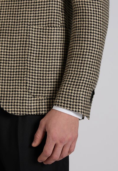 Nahaufnahme einer Hand neben dem Ärmel eines beige-schwarzen Blazers mit Hahnentrittmuster über einer weißen Hemdmanschette und schwarzen Hosen.