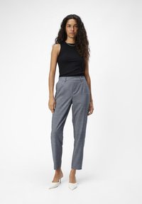 Schwarzes ärmelloses Top, graue karierte maßgeschneiderte Hose, glatte Textur, schlanker Schnitt, weiße spitze Absatzschuhe, silberne Kettenakzent am Ausschnitt.