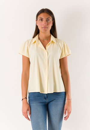 Giovane donna con lunghi capelli scuri che indossa una camicetta giallo pallido a maniche corte con bottoni e jeans blu, in piedi davanti a uno sfondo semplice.