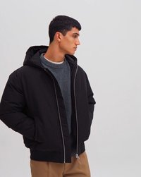 Junger Mann, der eine schwarze Kapuzenjacke über einem grauen Pullover und beigen Hosen trägt, mit den Händen in den Taschen stehend und seitlich blickend.