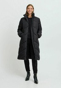 Abrigo largo negro tipo plumón con cuello alto, diseño acolchado y bolsillos laterales. Llevado sobre una blusa negra y pantalones ajustados con botines.