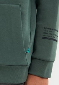 Zelená mikina s kapucí, přední kapsou, žebrovanými manžetami a texturovanou tkaninou. Obsahuje potištěný text a malou modrou etiketu na boku.