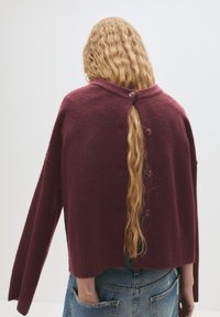 Maglione oversize bordeaux con collo alto, orlo a coste e dettagli con bottoni sul retro. Il materiale appare morbido con una finitura testurizzata.