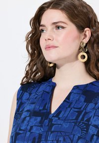 Camisetta senza maniche blu con motivi geometrici, dotata di scollatura a V. Grandi orecchini rotondi e texturizzati completano il look.