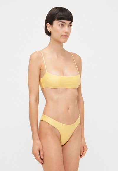 Dzeltena bikini komplekts ar minimālistisku dizainu, plānām siksnām un gludu tekstūru. Augšdaļai ir kvadrātveida dekoltē, bet apakšdaļas ir augsti griezti.
