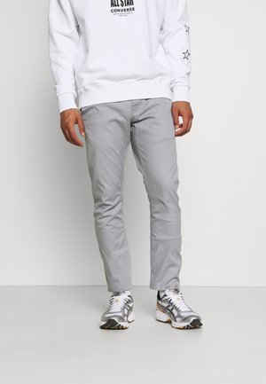 Mand iført lysegrå bukser, hvid Converse sweatshirt med stjernemønstre på ærmerne og hvide-grå sneakers stående indendørs.
