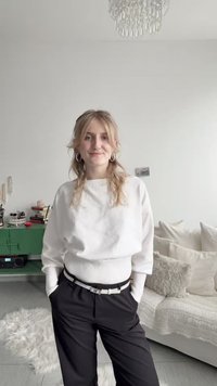 Νεαρή γυναίκα με ξανθά μαλλιά, φορώντας λευκό πουλόβερ και μαύρο παντελόνι, στέκεται σε ένα μοντέρνο σαλόνι με καναπέ, μαξιλάρια και πράσινη ντουλάπα.