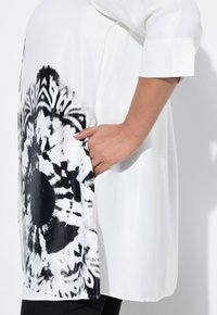 White tunic top with a bold black and white floral print on the side, featuring a relaxed fit and pocket detail. Smooth silk-like texture.