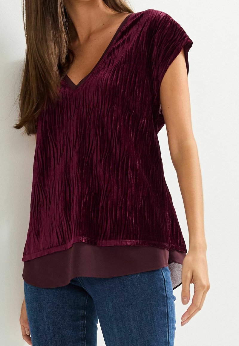 Femme portant un haut sans manches en velours texturé bordeaux avec un ourlet à couches, associé à un jean bleu sur un fond uni.