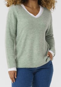 Jasnozielony sweter z prążkowanym wzorem, z dekoltem w kształcie litery V i długimi rękawami, zestawiony z białą koszulką. Sweter ma miękką teksturę i luźny krój.