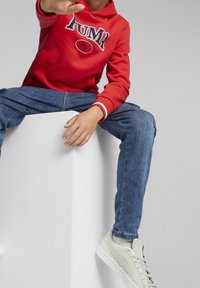 Sweatshirt rouge avec le logo "PUMA", jeans bleus et baskets gris clair. Le sweatshirt possède une poche kangourou et des accents blancs sur les manches.