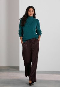 Pulover teal cu guler tip tunică, cu manșete crem și decolteu în formă de V, asortat cu pantaloni satinați de culoare maro închis, cu croi larg și tocuri negre.