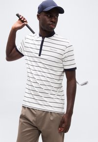 Άνδρας με μπλε σκουφί, λευκό κοντομάνικο πόλο με ρίγες και μπεζ παντελόνι κρατά γκολφ ρόπαλο στον ώμο του, κοιτάζοντας πλάγια σε μονόχρωμο φόντο.