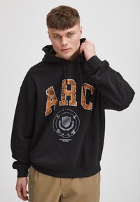 Sudadera con capucha negra hecha de un tejido suave, con letras en relieve naranjas "ARC" y un logotipo gráfico blanco en el centro.