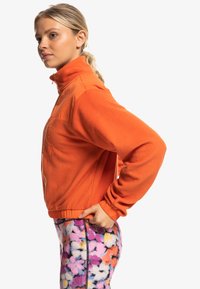 Pomarańczowy polarowy sweter z wysokim kołnierzem, z przednią kieszenią i elastycznym pasem; zestawiony z leginsami w kwiatowy wzór w żywych kolorach.
