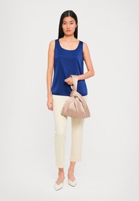 Woman wearing a sleeveless blue top and cream pants, holding a beige knotted satin handbag, standing against a plain background.