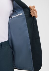 Man in een teal kleurig pak toont de gladde satijnen voering met een binnenzak van het jasje tegen een witte achtergrond.