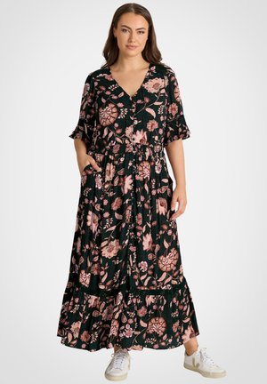 Kobieta ubrana w czarną kwiecistą sukienkę midi z różowymi kwiatami, krótkimi rękawami, kieszeniami i białe sportowe trampki, stojąca na białym tle.