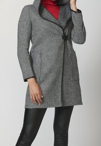 Cappotto a scacchi bianco e nero, con un grande colletto, due tasche laterali e una silhouette aderente. Indossato sopra un dolcevita rosso.