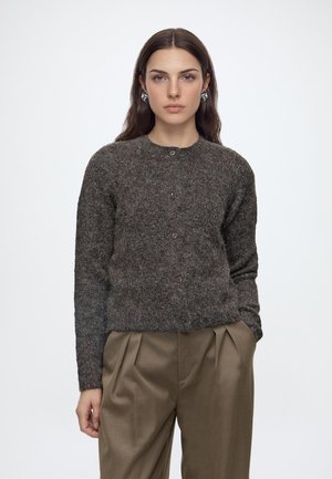 Jeune femme aux longs cheveux bruns portant un cardigan gris boutonné et un pantalon plissé marron, debout avec une main dans la poche sur fond blanc.