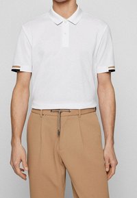 Polo shirt blanc avec un col classique, présentant des accents rayés noirs et beige sur les manches, associé à un pantalon texturé beige.